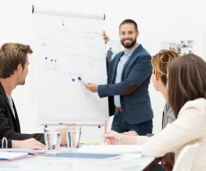 Ein Mann präsentiert Inhalte an einem Flipchart vor mehreren Teilnehmenden in einem hellen Besprechungsraum.