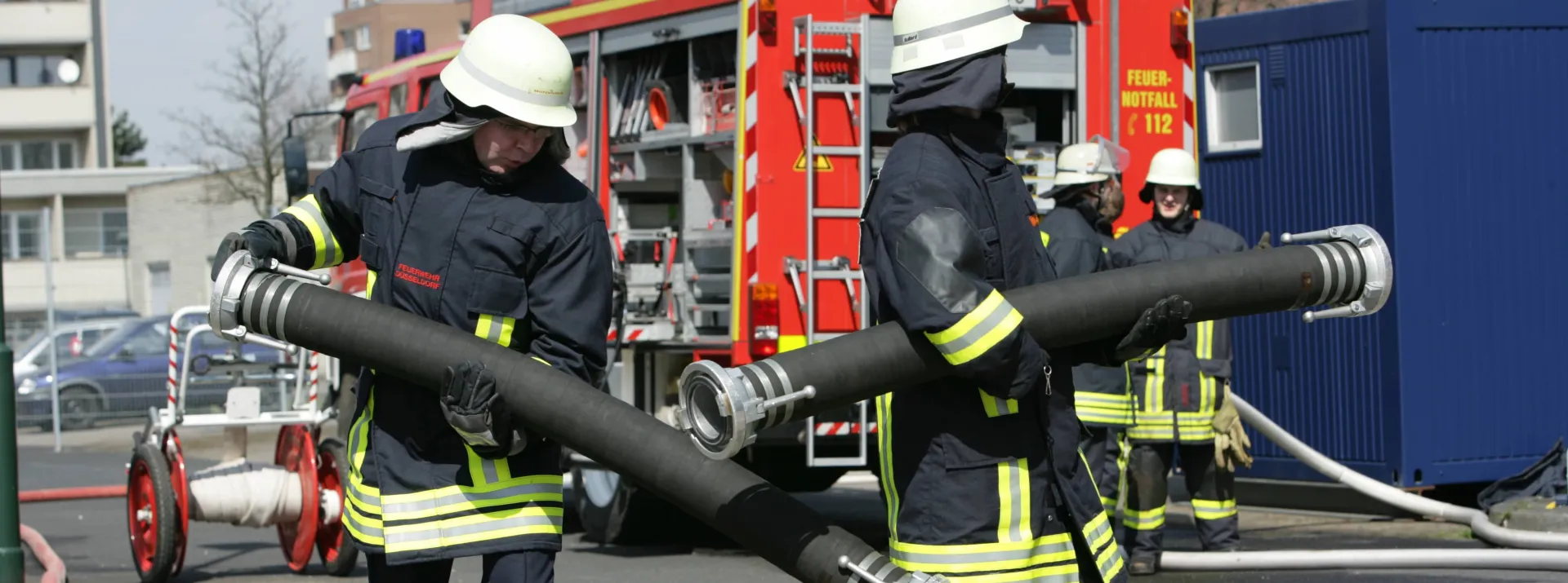 Feuerwehrleute in Schutzkleidung tragen eine große Schlauchleitung vor einem Feuerwehrfahrzeug während einer Einsatzübung.